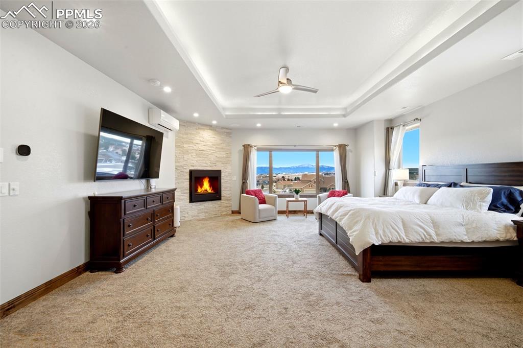 Image 20 of 48: Bedroom with a fireplace, a ceiling fan, light carpet, a tray ceiling, and