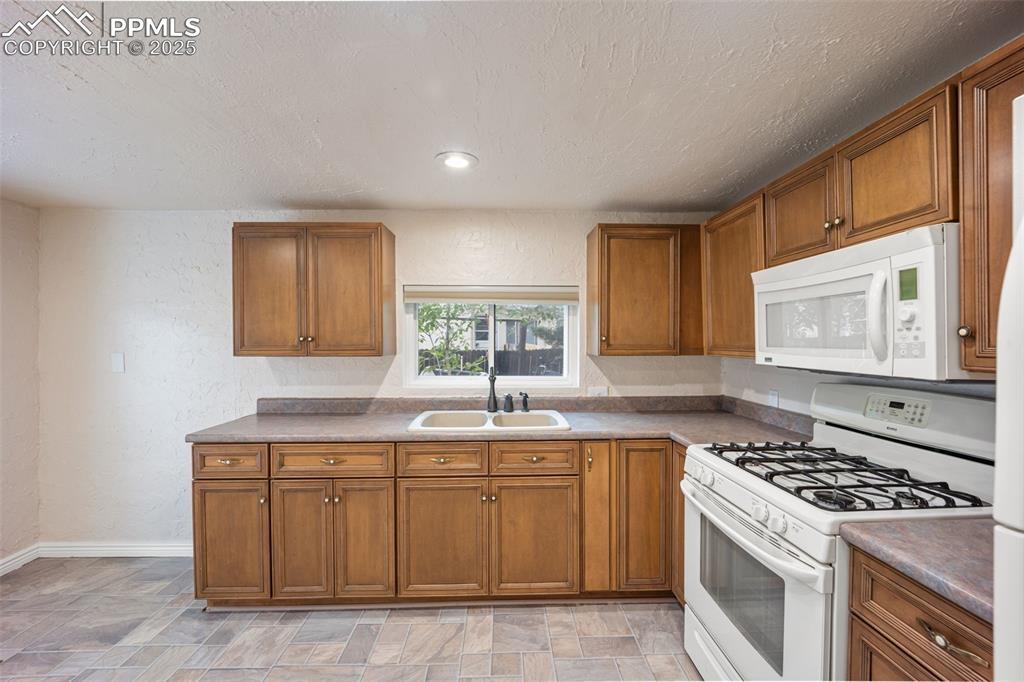 Image 12 of 31: Kitchen with white appliances, a textured wall, brown cabinets, stone finis