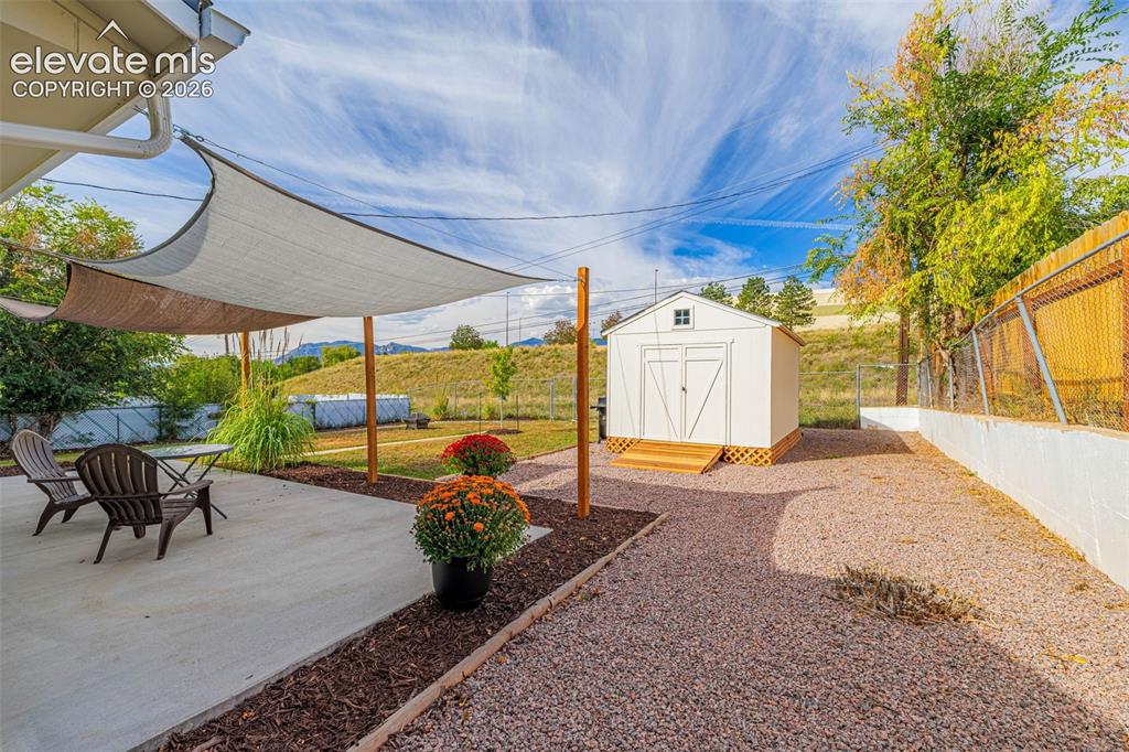 Image 19 of 27: Concrete patio area, shade sail, gravel landscaping, white storage shed wit