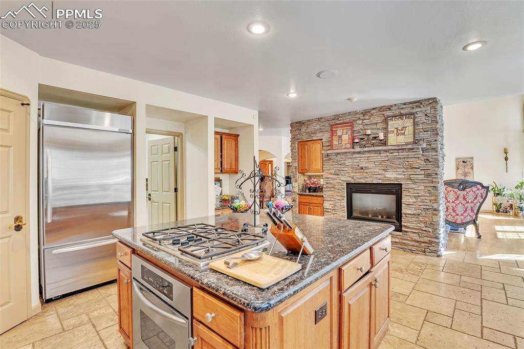 Image 9 of 45: Kitchen with dark stone counters, a stone fireplace, an island with sink, s