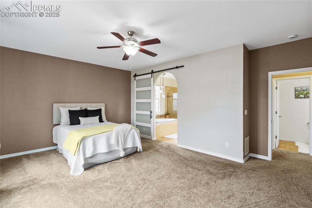 Image 21 of 36: Bedroom with a barn door, carpet, ceiling fan, and ensuite bath