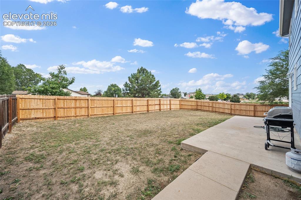 Image 28 of 41: Wide view of the fenced backyard with concrete patio, mature trees nearby, 