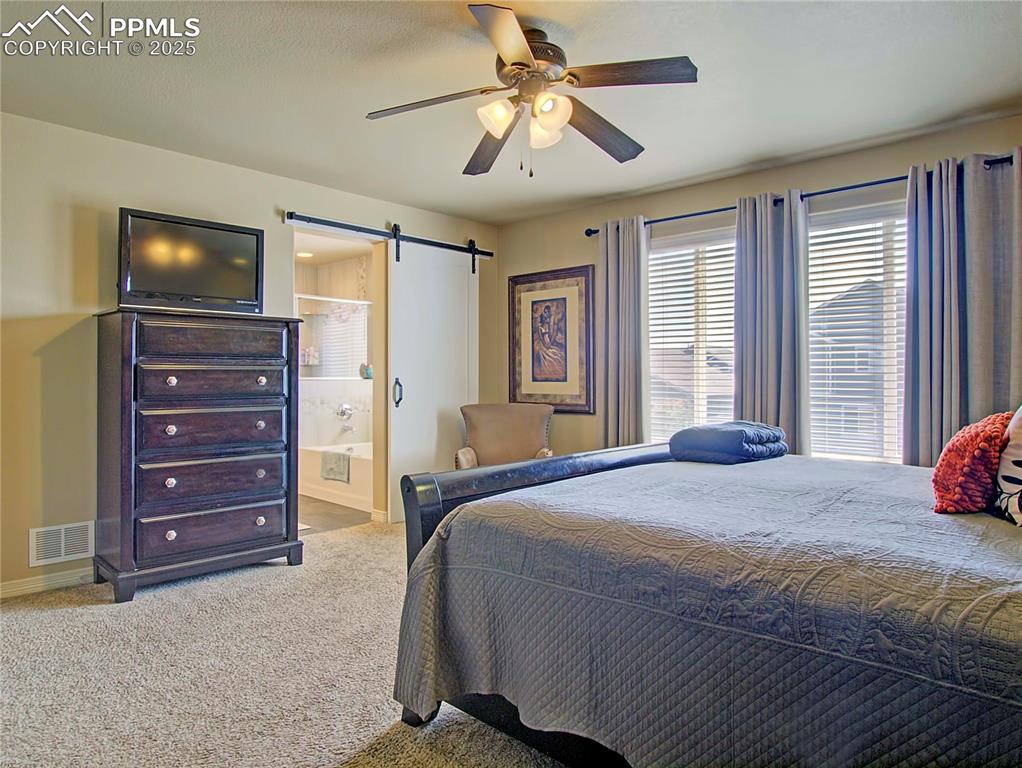 Image 24 of 30: Carpeted bedroom featuring a barn door, a ceiling fan, and ensuite bath