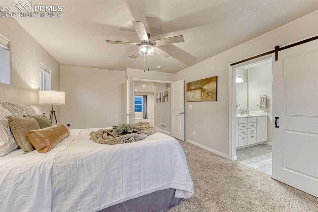 Image 34 of 50: Bedroom with a barn door, light colored carpet, ceiling fan, and ensuite ba