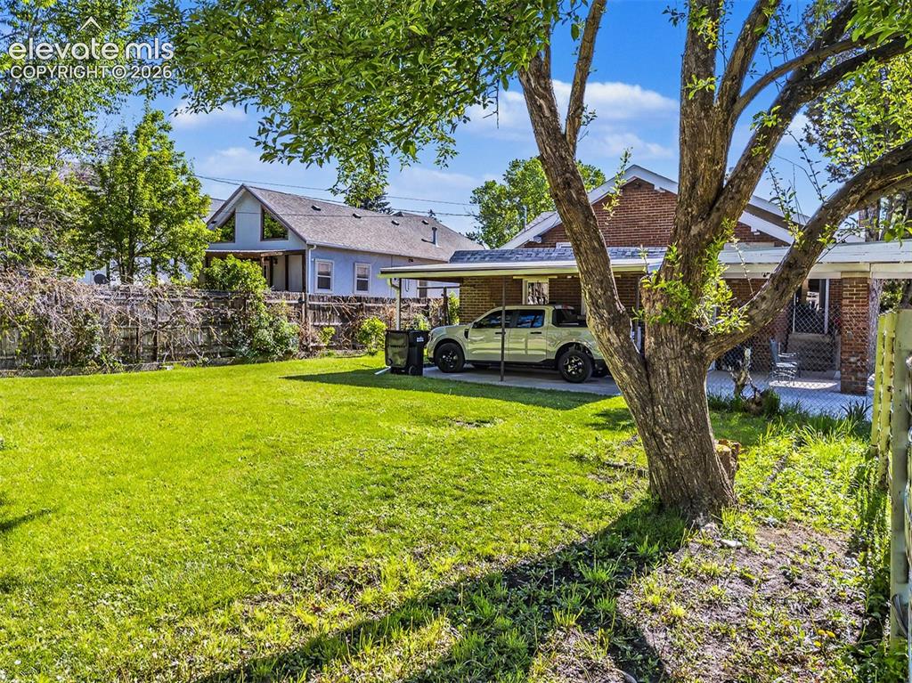 Image 19 of 27: Spacious lawn with a mature tree, featuring a brick exterior and a covered