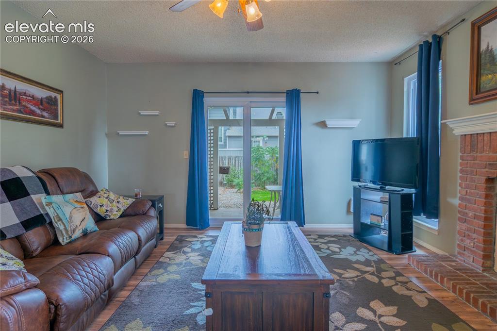 Image 12 of 42: Living area with wood-finish flooring, a brick fireplace, and a ceiling fan