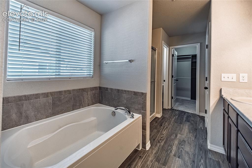 Image 21 of 35: White soaking tub with tiled surround, wood-finish flooring, and a vanity w