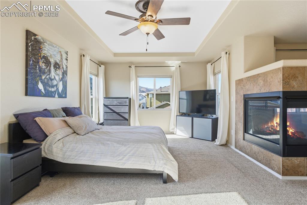Image 16 of 42: Carpeted bedroom featuring a raised ceiling, a ceiling fan, and a fireplace