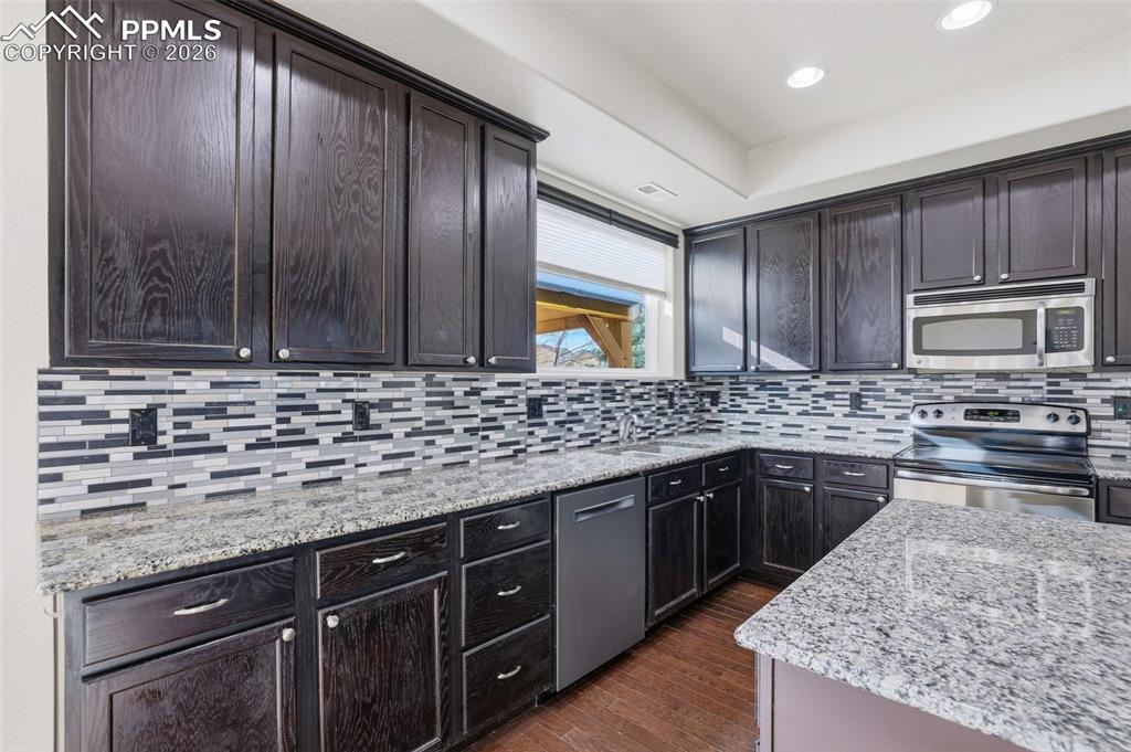 Image 18 of 39: Kitchen with stainless steel appliances, light stone counters, dark wood-ty