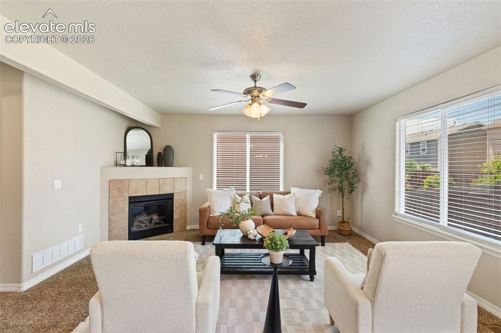 Image 19 of 35: Living area with a tiled fireplace, large windows with blinds, and a ceilin