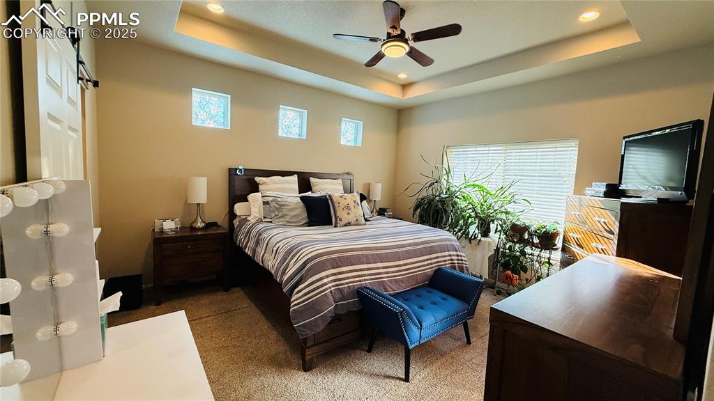 Image 13 of 28: Bedroom featuring a tray ceiling, multiple windows, ceiling fan, a barn doo
