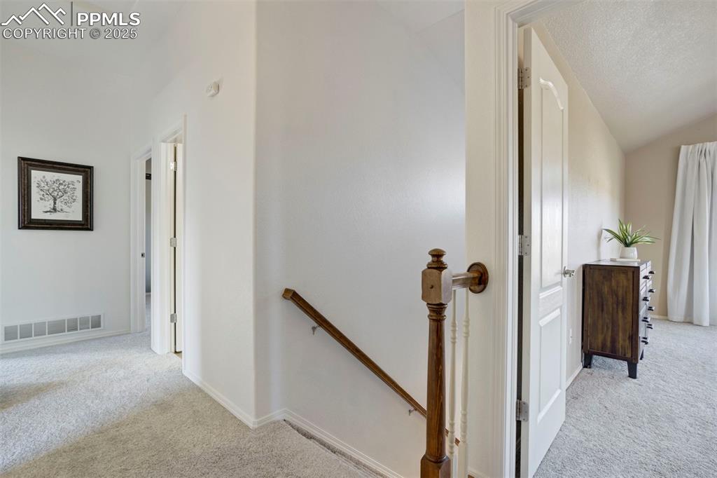Image 31 of 50: Corridor with light carpet, an upstairs landing, lofted ceiling, and a text