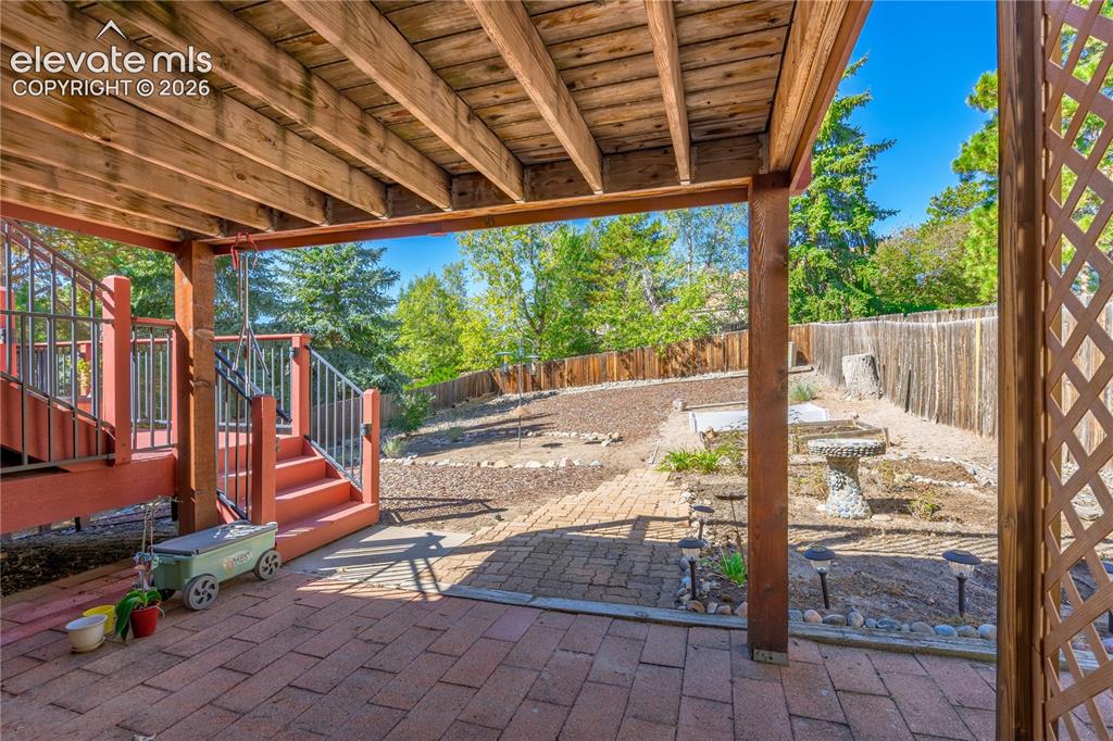 Image 28 of 43: Covered patio with brick pavers, wood deck with stairs, wood lattice, and f