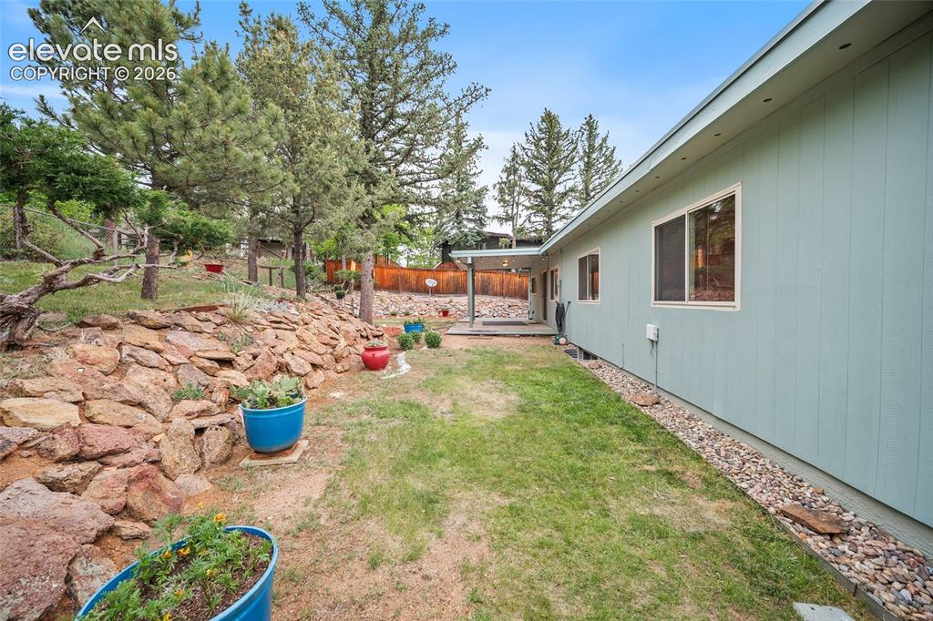 Image 37 of 50: Expansive backyard featuring a stone retaining wall, mature trees, a covere