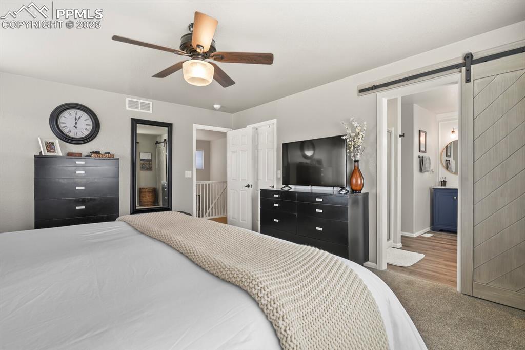 Image 30 of 40: Carpeted bedroom with a barn door, ceiling fan, and ensuite bath