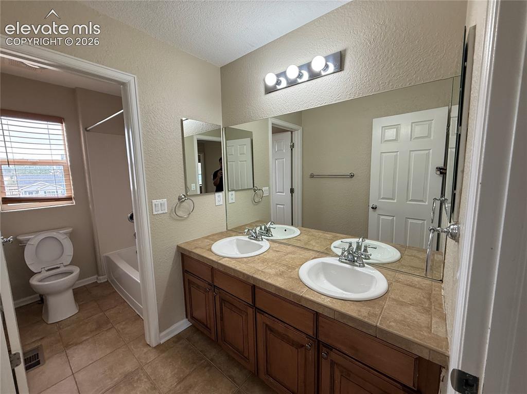 Image 17 of 28: Dual vanity with wood-finish cabinetry and tile countertop, large wall-moun