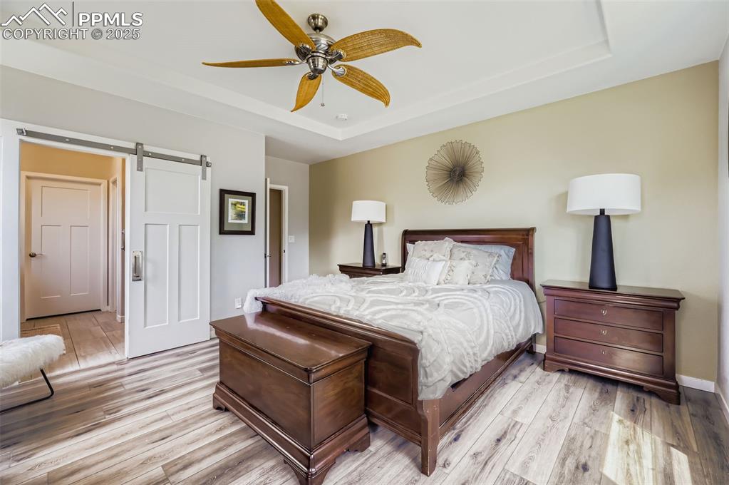 Image 21 of 50: Bedroom featuring a barn door, a raised ceiling, ceiling fan, and light woo