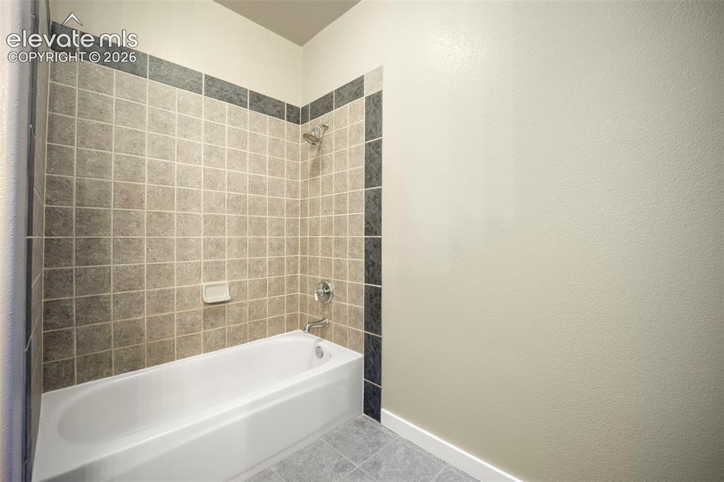 Image 36 of 40: Bathtub with a tiled surround featuring light-toned square tiles and a dark