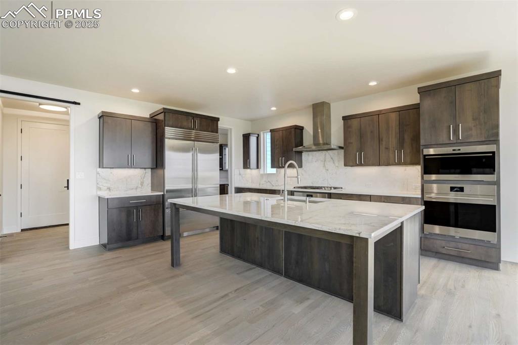Image 8 of 34: Kitchen featuring dark brown cabinets, backsplash, an island with sink, app
