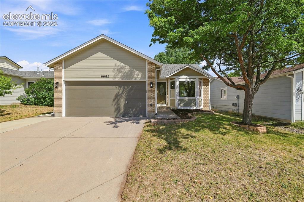 Caption: Two-car garage with concrete driveway, bay window, landscaped entry, and mature tree