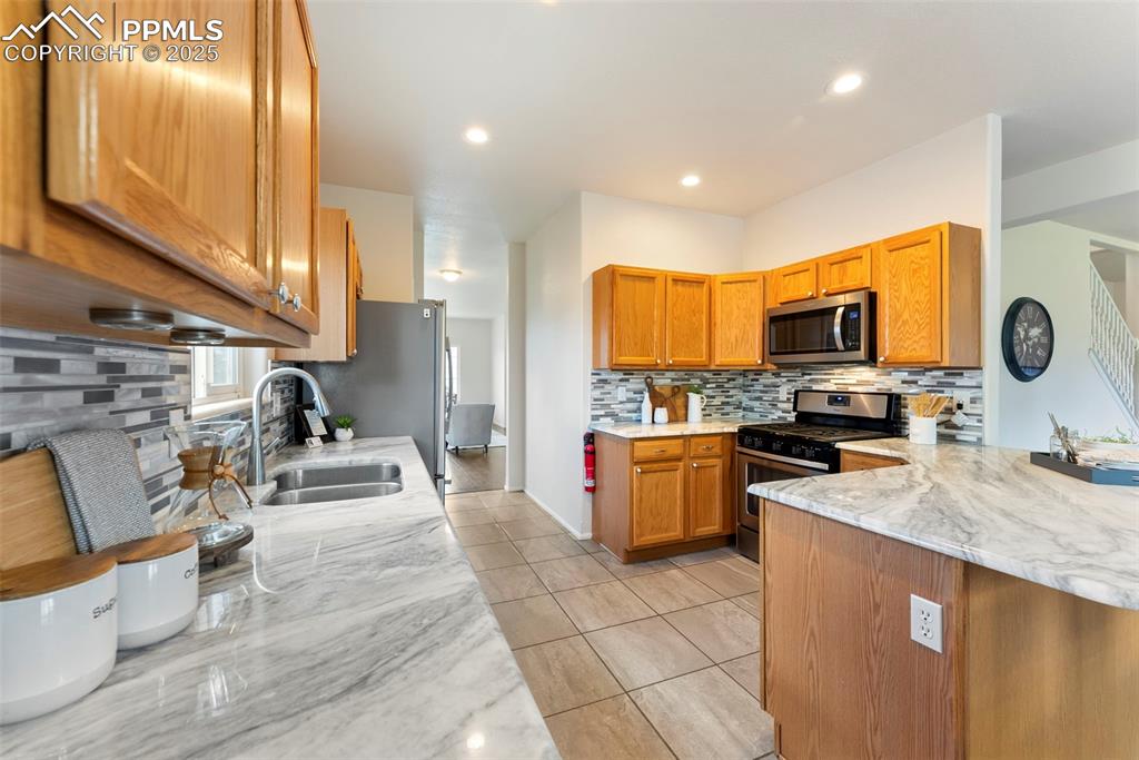 Image 6 of 50: Kitchen featuring backsplash, brown cabinetry, light stone countertops, sta