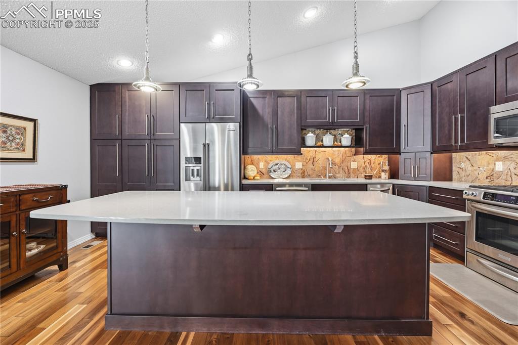 Image 15 of 50: Kitchen featuring lofted ceiling, dark brown cabinetry, pendant lighting, a