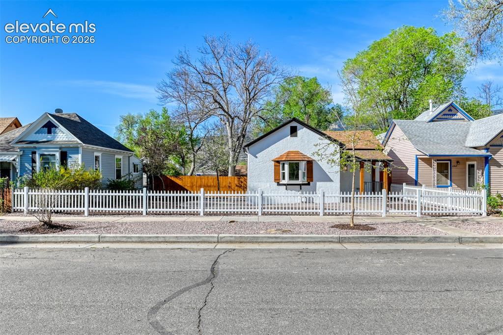 Image 2 of 47: White picket fence framing a stucco exterior with a prominent bay window fe