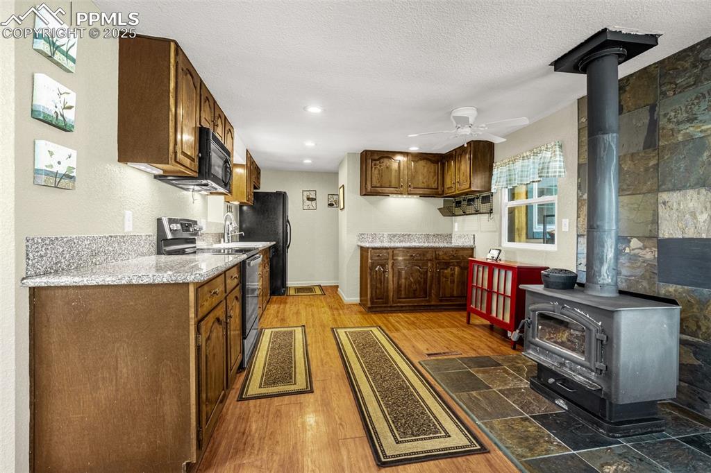 Image 6 of 31: Kitchen with black appliances, a wood stove, a ceiling fan, light wood fini