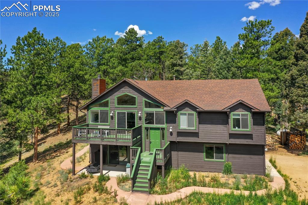 Caption: View of front of house with a wooden deck, stairway, a chimney, a patio, and roof with shingles