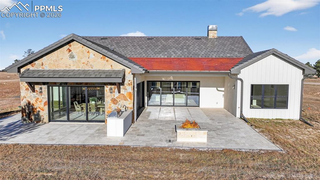 Image 8 of 49: Back of house with stone siding, a chimney, a patio area, and roof with shi