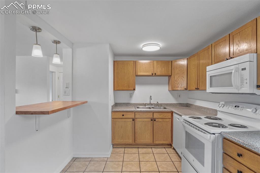 Image 9 of 17: Kitchen with white appliances, pendant lighting, brown cabinetry, a breakfa