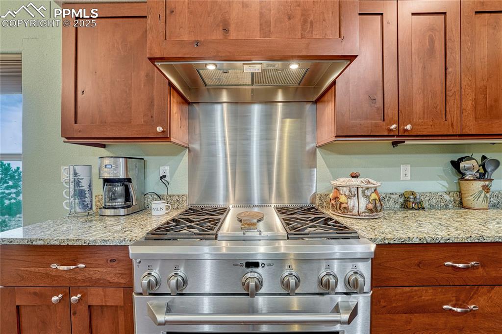 Image 13 of 43: Kitchen featuring high end range, brown cabinetry, light stone countertops,
