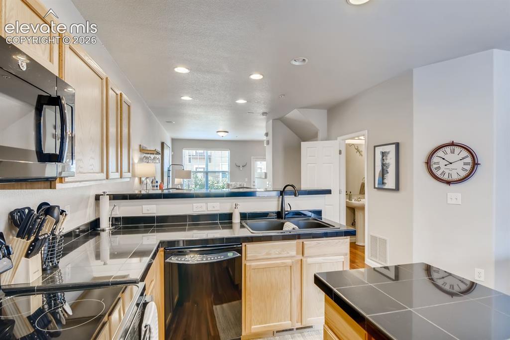 Image 10 of 23: Well-appointed kitchen featuring light wood cabinetry, black tile counterto
