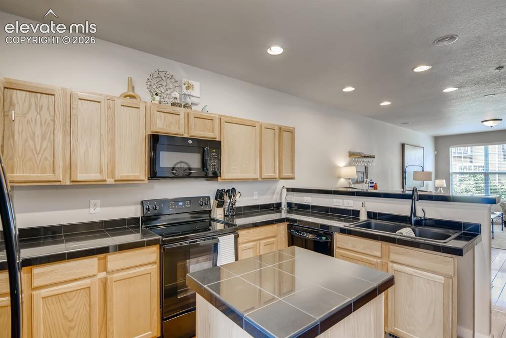 Image 9 of 23: Kitchen featuring light wood cabinetry, black tile countertops, black appli