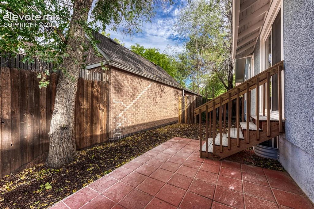 Image 36 of 44: Patio area featuring red paver tiles, wooden privacy fencing, mature trees,
