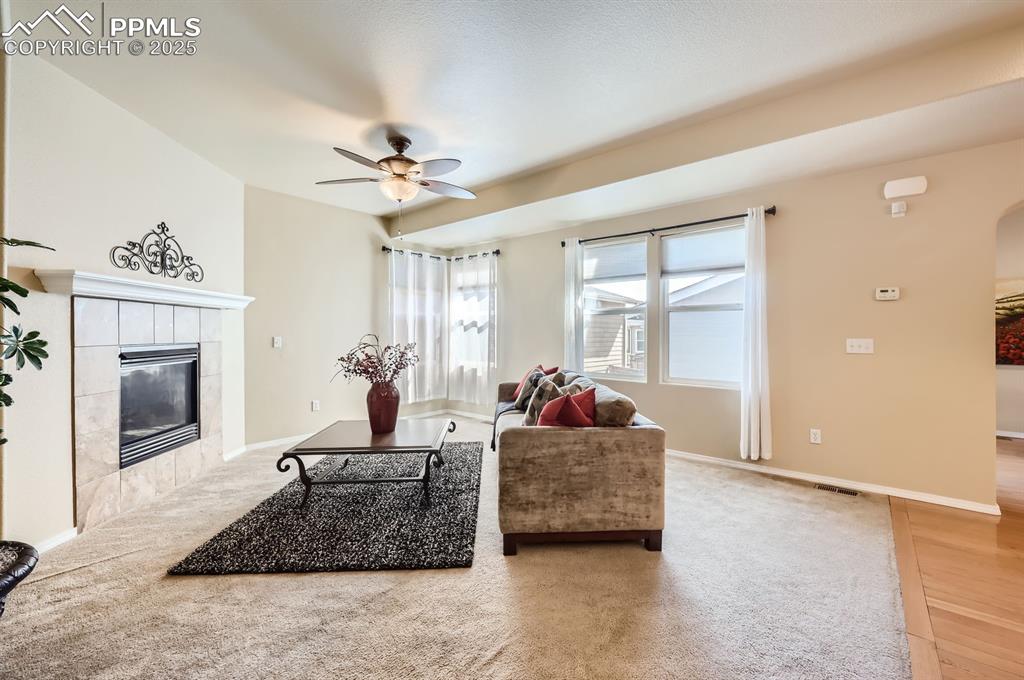 Image 11 of 34: Living area with ceiling fan, a tiled fireplace, light colored carpet, arch