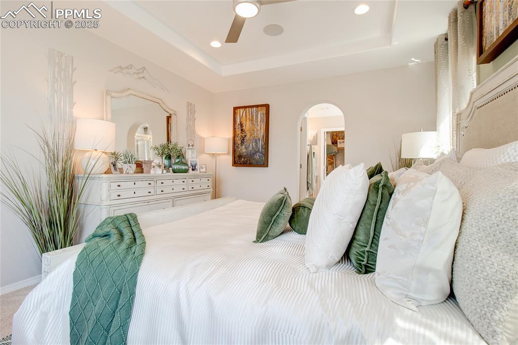 Image 17 of 23: Carpeted bedroom with arched walkways, a tray ceiling, a ceiling fan, and r