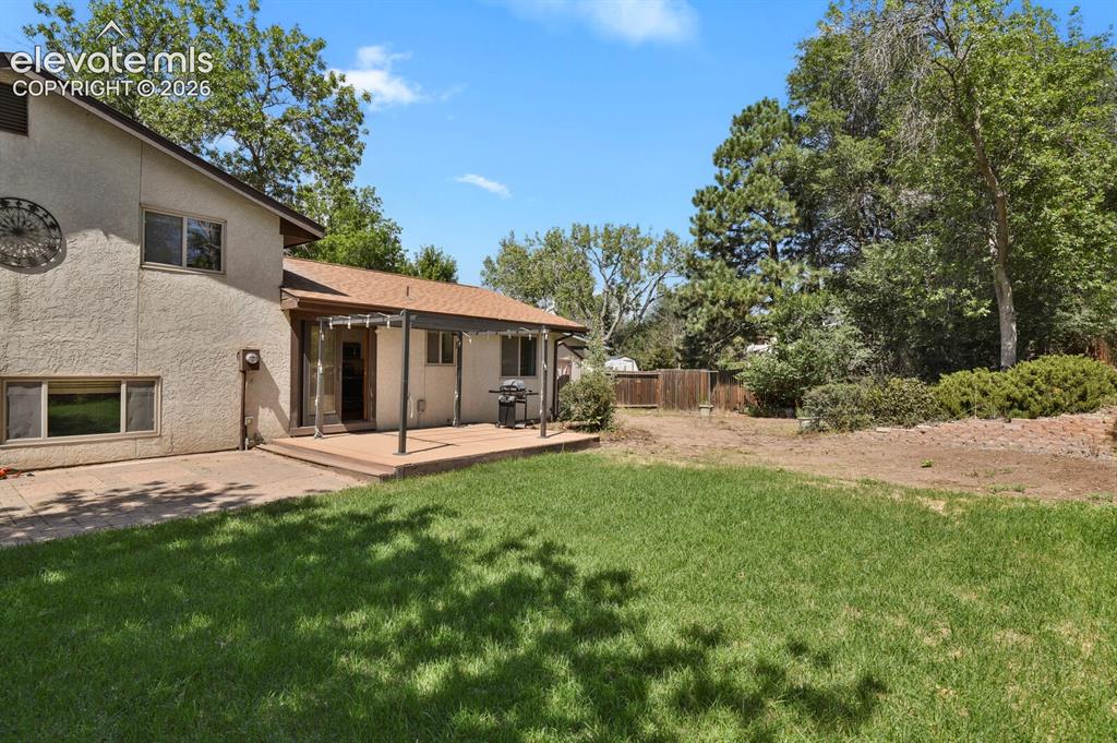 Image 29 of 34: Expansive grassy yard with a covered patio, multiple windows, and mature tr