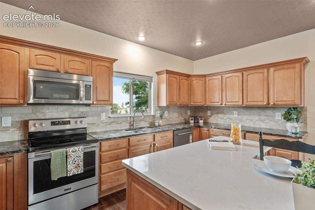 Image 14 of 49: Kitchen featuring wood cabinetry, stainless steel appliances, dark stone co