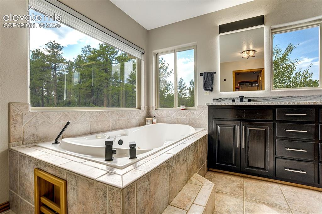 Image 23 of 40: Soaking tub with tile surround, expansive windows, dark wood-finish vanity 