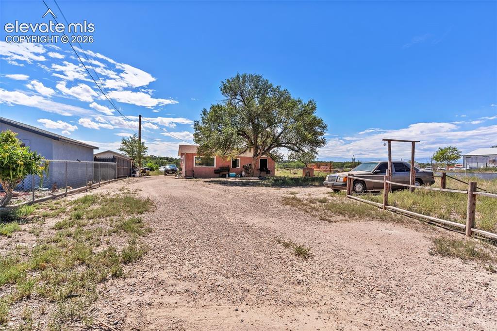Image 23 of 29: Gravel driveway with a wooden fence entrance, mature tree, and a single-sto