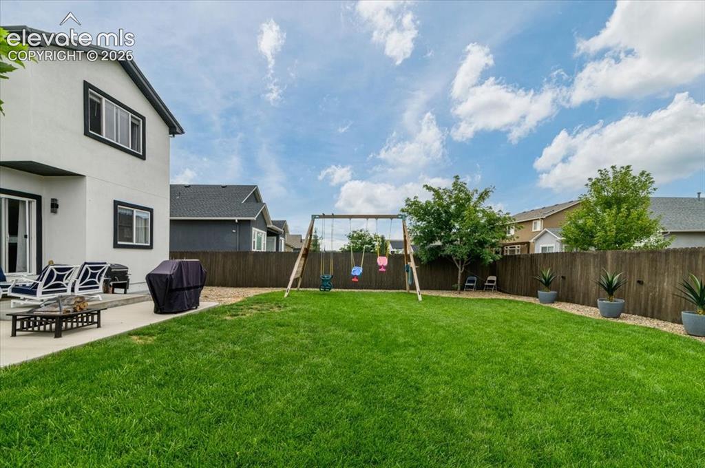 Image 34 of 39: Spacious fenced backyard featuring a swing set, mature trees, a concrete pa