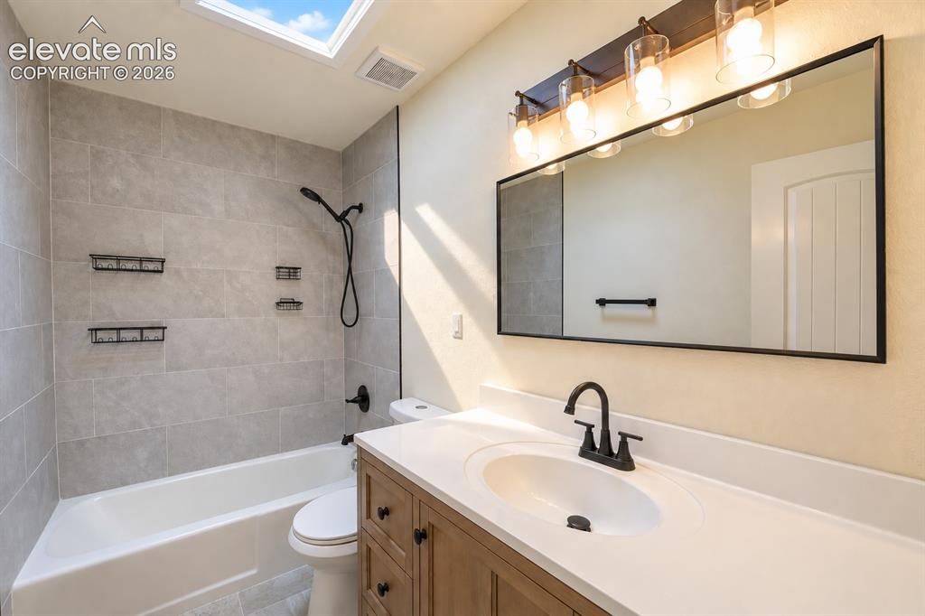 Image 9 of 29: White bathtub with light gray tile surround, wood-finish vanity with counte