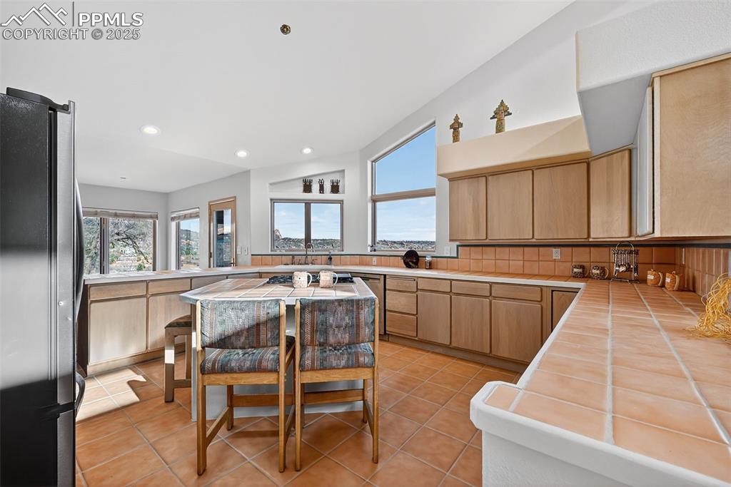 Image 15 of 50: Kitchen with tile counters, freestanding refrigerator, light brown cabinetr