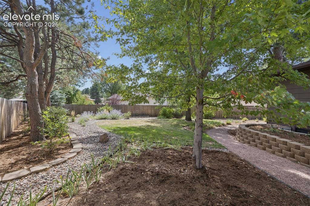 Image 32 of 36: Backyard featuring a stone-retaining wall, gravel pathway, mature trees, ti