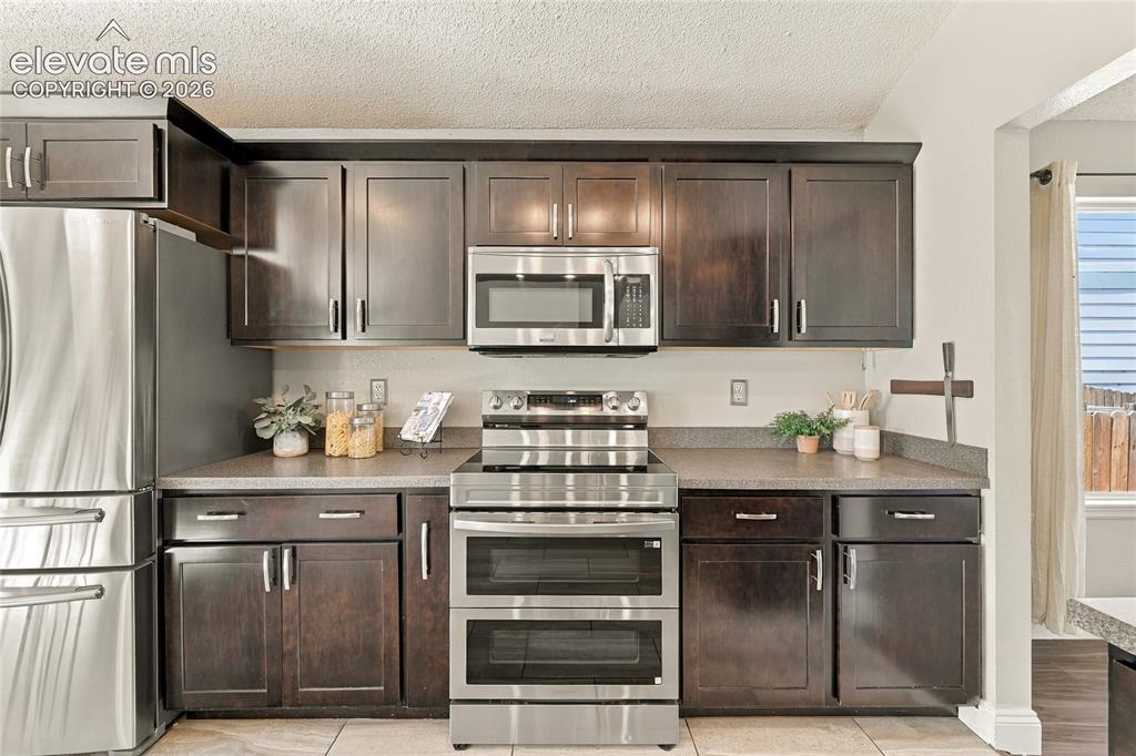 Image 14 of 45: Kitchen featuring dark wood cabinetry, stainless steel appliances, stone-fi
