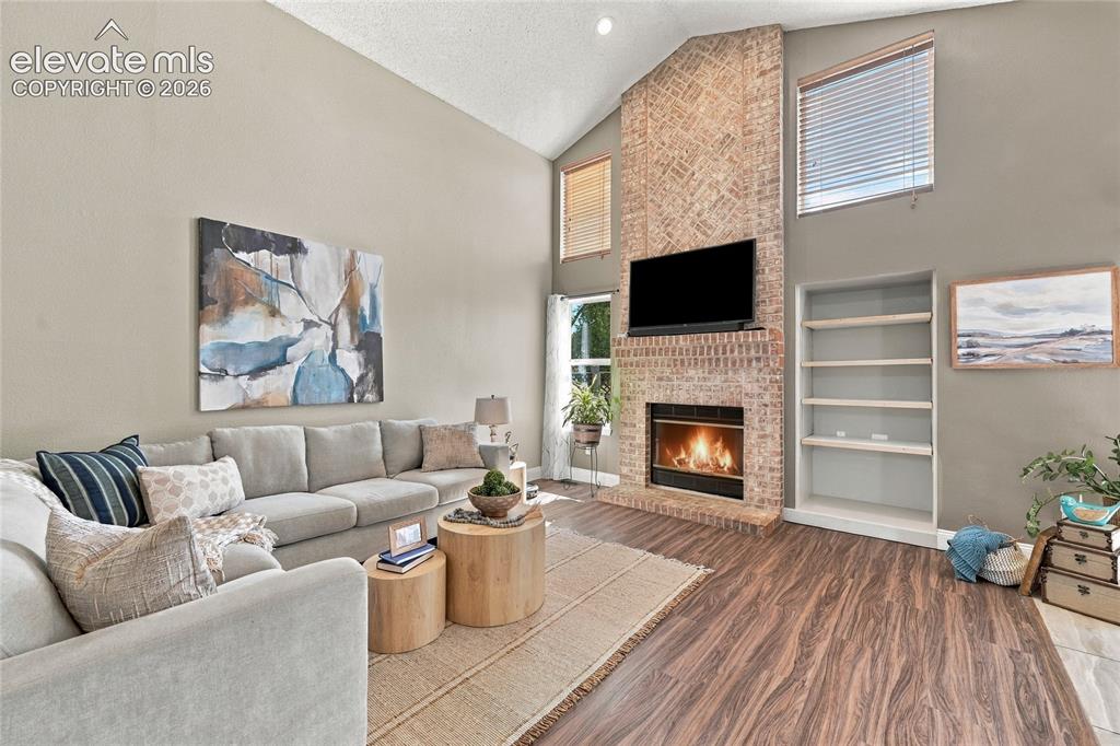 Image 7 of 45: Vaulted ceiling, brick-faced fireplace, built-in shelving, wood look vinyl