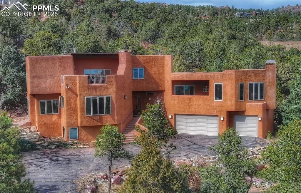 Image 2 of 20: Pueblo-style house featuring aphalt driveway, a wooded view, stairs, a gara