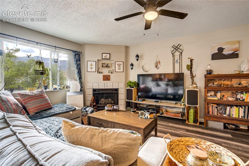 Image 31 of 50: Corner fireplace with tile surround, wood-finish flooring, and a ceiling fa