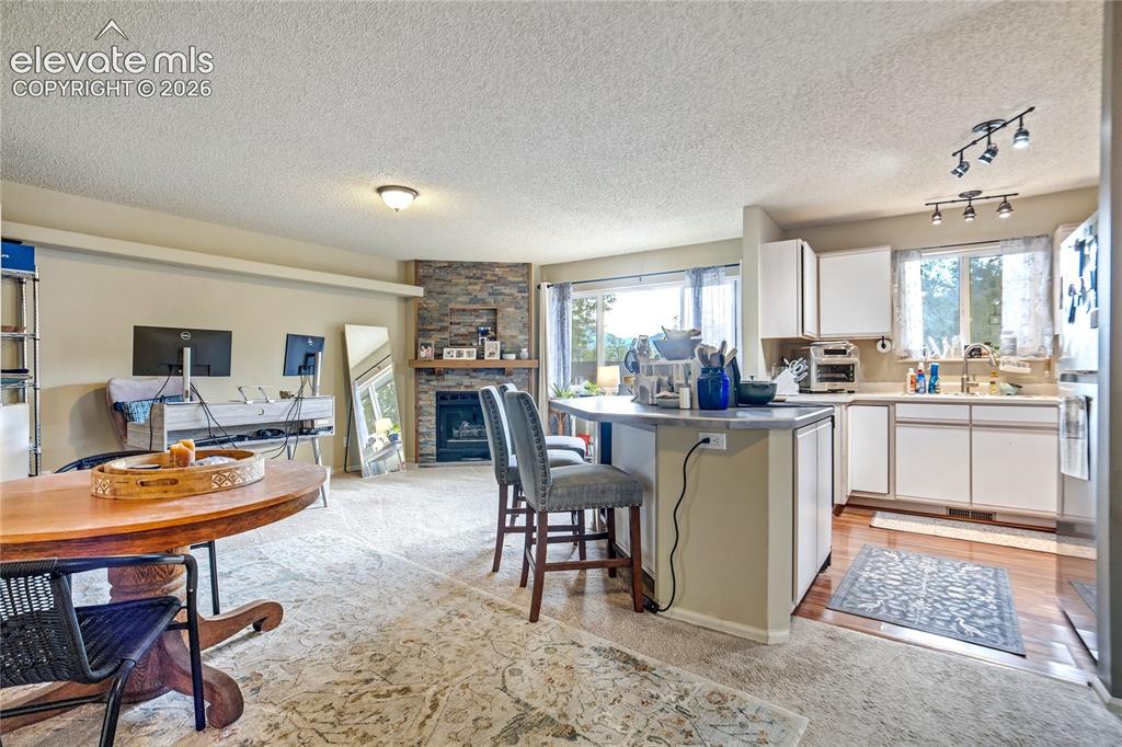 Image 8 of 50: Living area with a stone-clad corner fireplace, an open kitchen with white 